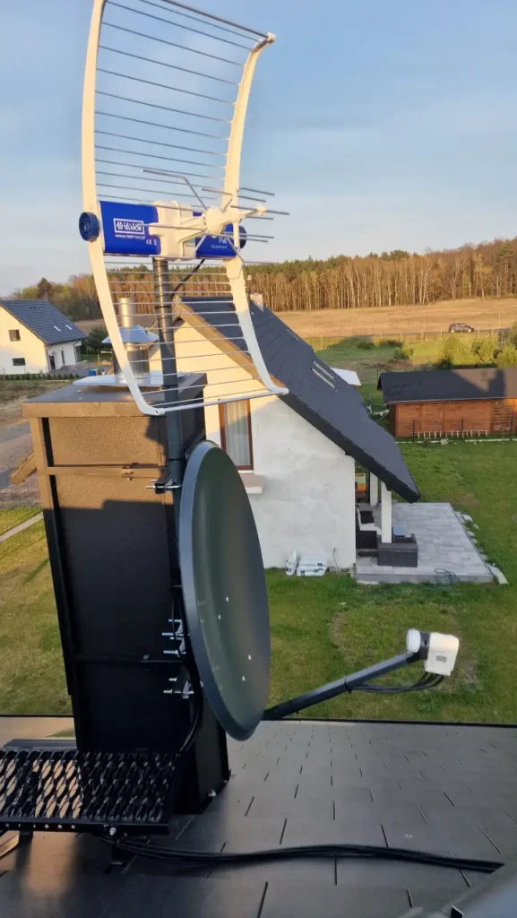 Czarna antena satelitarna na kominie, nad nią antena kierunkowa DVB-T2. W tle doki i las
