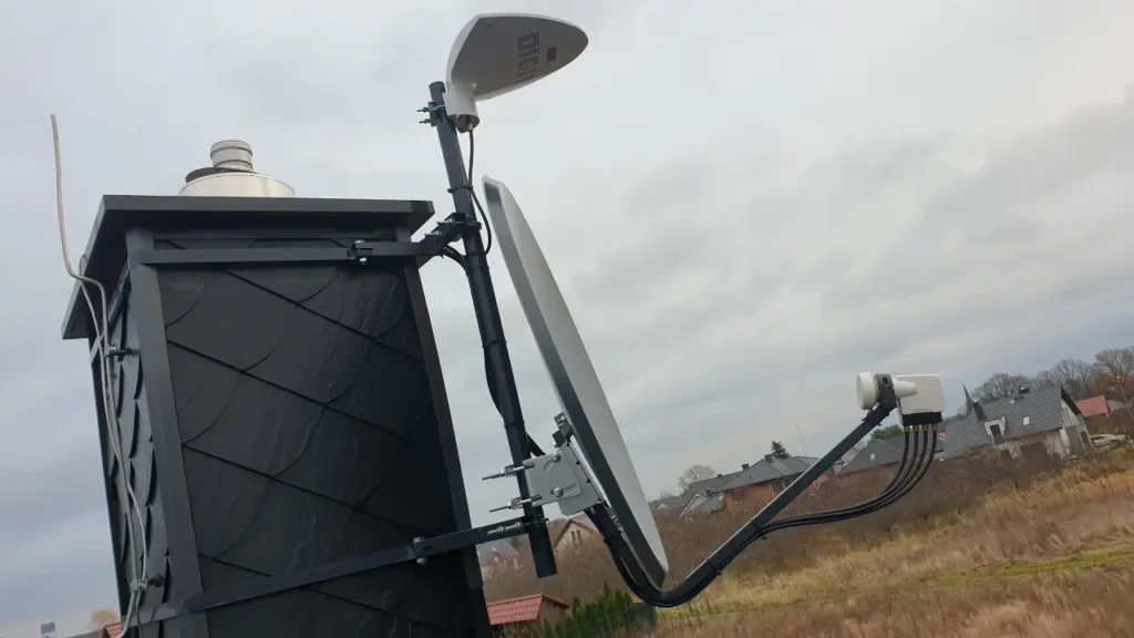 Jasna antena satelitarna zamontowana na kominie. W tle zachmurzone niebo i domki jednorodzinne
