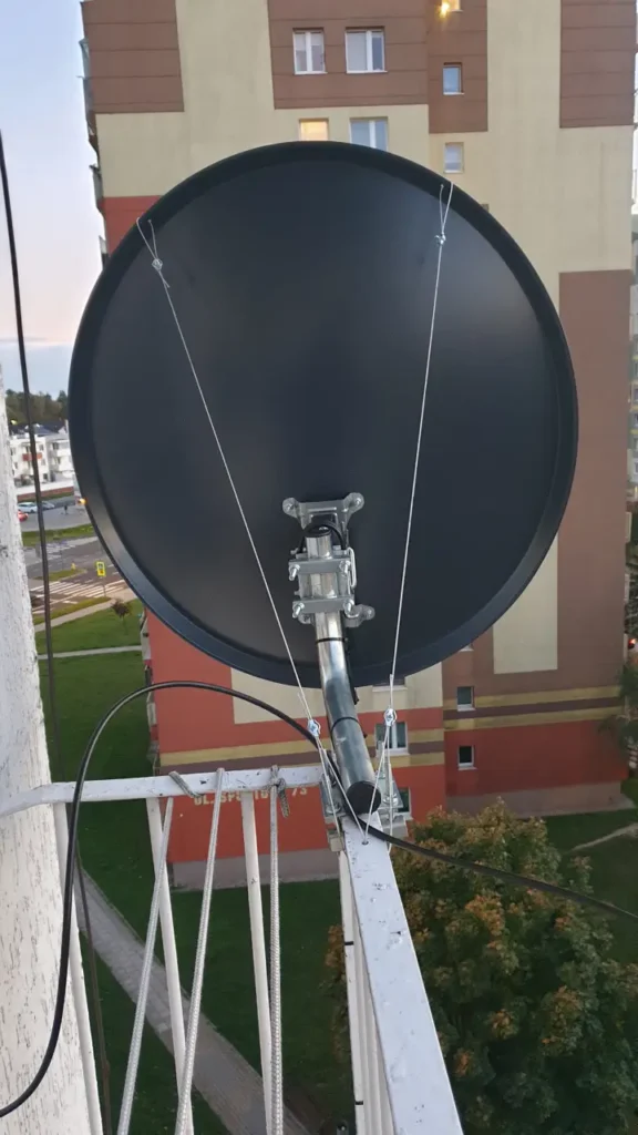Czarna antena satelitarna zamontowana na balkonie z dodatkowymi linkami wzmacniającymi montowanie. W tle budynek wielorodzinny