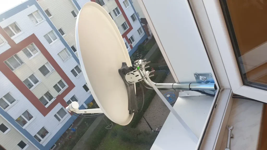 Biała antena satelitarna zamontowana w oknie bloku wielorodzinnego na wysokim piętrze. W tle widok na różnokolorowy blok wielorodzinny