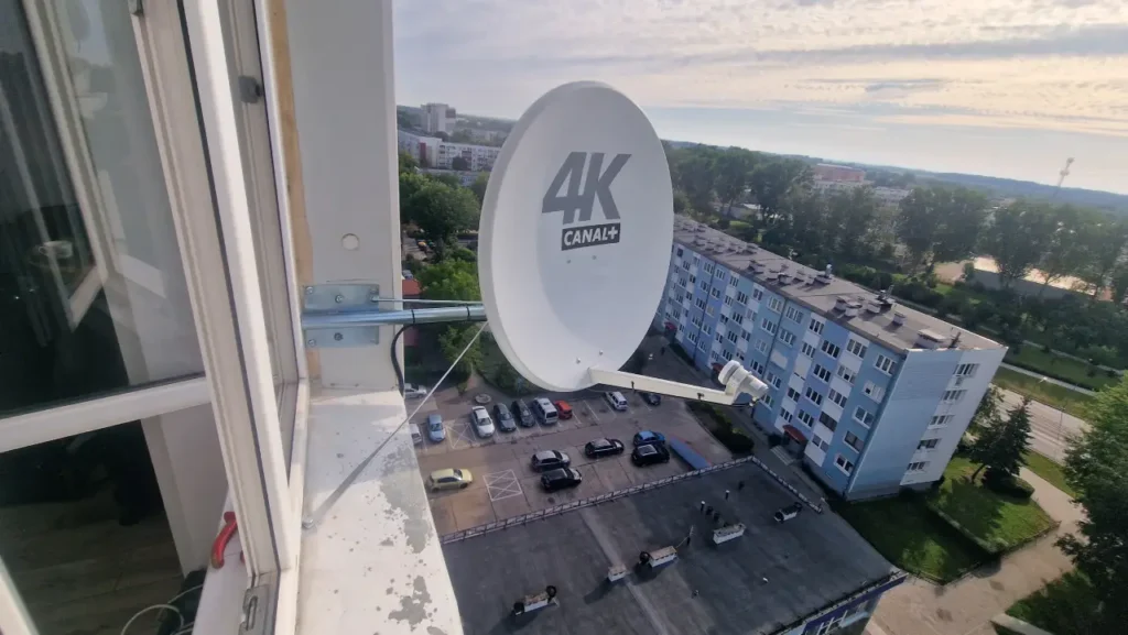 Biała antena satelitarna zamontowana w oknie bloku wielorodzinnego na wysokim piętrze. W tle widok na różnokolorowy blok wielorodzinny oraz parking