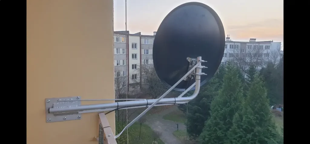 Czarna antena satelitarna na narożniku elewacji na wysokiej kondygnacji. Widok na ciąg bloku wielorodzinnego. W dole wysokie drzewa iglaste