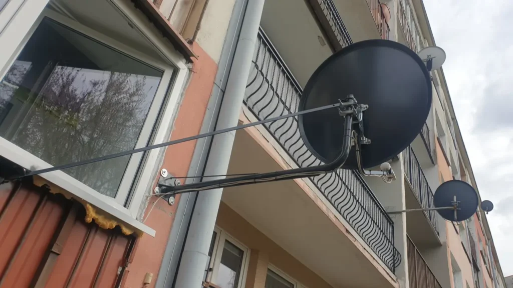 Czarna antena satelitarna zamontowana na elewacji przy oknie. Na tej samej elewacji jeszcze kilka anten satelitarnych