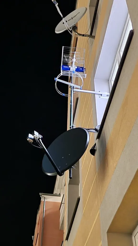 Czarna antena satelitarna, nad nią antena kierunkowa DVB T2 u jeszcze wyżej jasna antena satelitarna, wszystko to na żółtej elewacji w tle noc
