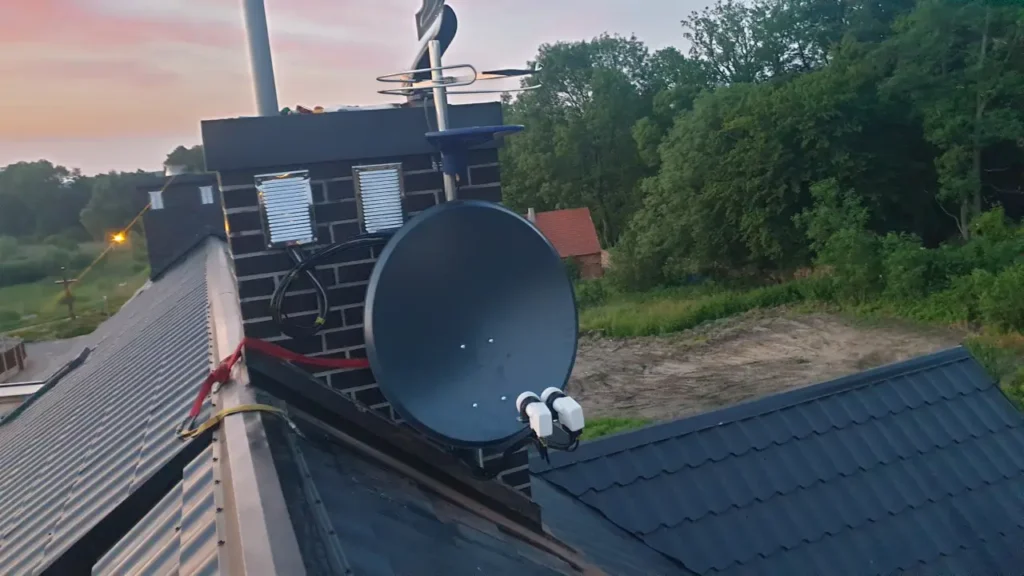 Czarna antena satelitarna z dwoma konwerterami. Antena zamontowana na kominie. W tle zielone drzewa 