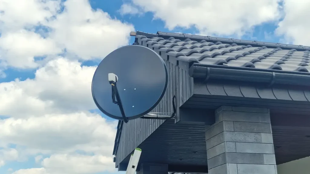 Czarna antena satelitarna pod czarnym dachem. W tle błękitne niebo z białymi obłokami